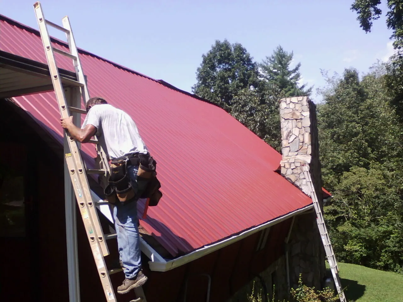 Roofer on a ladder beside a red metal roof and stone chimney — Soffit & Fascia Repair by Roofing Brothers in Egan
